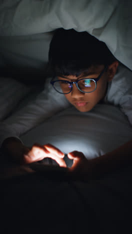 Vertical-Video-Close-Up-Of-Young-Boy-In-Bedroom-At-Home-Using-Mobile-Phone-To-Text-Message-Under-Covers-Or-Duvet-At-Night-6